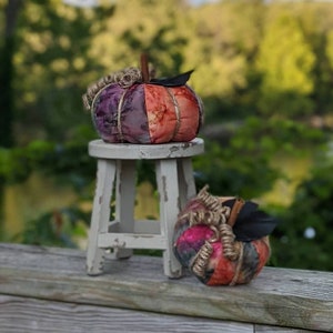 May include: Two decorative fabric pumpkins in shades of purple, orange, and red, with twine accents and faux leaves. One pumpkin sits on a small, distressed white stool, the other on a wooden railing. Fall decor.