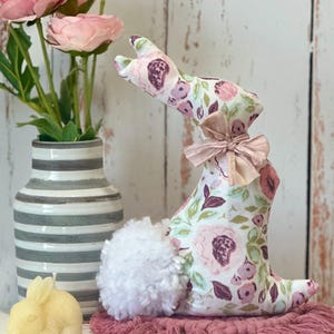 May include: A floral patterned bunny rabbit with a white pom-pom tail. The bunny is sitting on a pink fringed rug. There is a vase of pink flowers and a yellow bunny candle in the background.