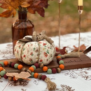 May include: A decorative fabric pumpkin with a floral pattern, twine stem, and a wooden bead garland in fall colors. The pumpkin sits on a wooden board with the word "thankful" carved into it, alongside a brown glass vase with autumn leaves.