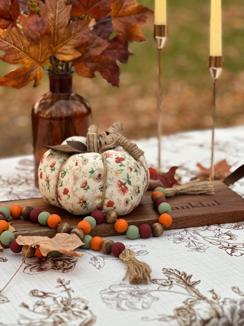 May include: A decorative fabric pumpkin with a floral pattern, twine stem, and a wooden bead garland in fall colors. The pumpkin sits on a wooden board with the word "thankful" carved into it, alongside a brown glass vase with autumn leaves.