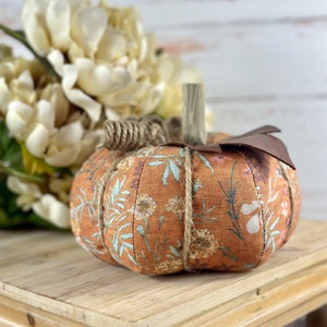May include: A fabric pumpkin with an orange floral pattern, a wooden stem, and a brown leaf. The pumpkin is tied with twine and sits on a wooden surface. Cream-coloured flowers are in the background.