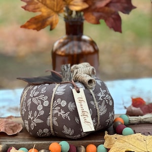 May include: A brown fabric pumpkin with a white floral pattern and a wooden stem. A small wooden tag with the word "thankful" hangs from the stem. The pumpkin is decorated with twine and a dark brown leaf.