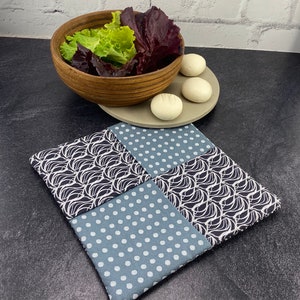 May include: A square, quilted hot pad with a patchwork design. Two squares feature a navy blue and white wave pattern, and two squares have a light blue polka dot pattern. A wooden bowl of lettuce and a plate with three white buns are in the background.
