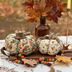 May include: Two fabric pumpkins with floral patterns sit on a wooden board, alongside a garland of colorful beads and autumn leaves. A brown vase holds fall foliage, and a candle holder is in the background. The pumpkins have twine stems.