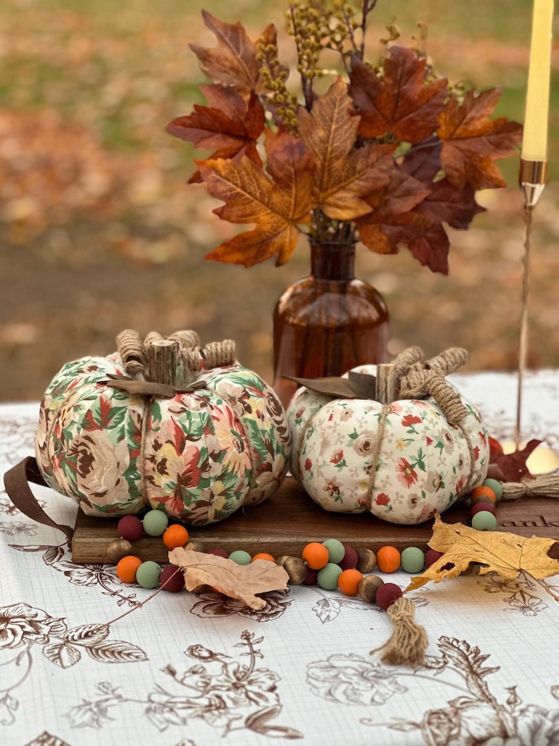 May include: Two fabric pumpkins with floral patterns sit on a wooden board, alongside a garland of colorful beads and autumn leaves. A brown vase holds fall foliage, and a candle holder is in the background. The pumpkins have twine stems.