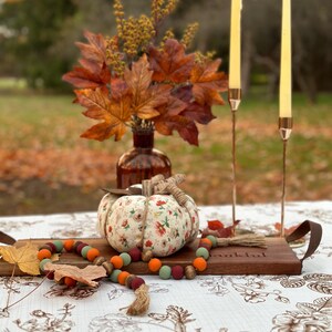 May include: A fall-themed table setting featuring a fabric pumpkin with floral patterns, a wooden tray, and a beaded garland. Two slender candles stand in copper holders, and a vase of autumn leaves adds to the seasonal decor.
