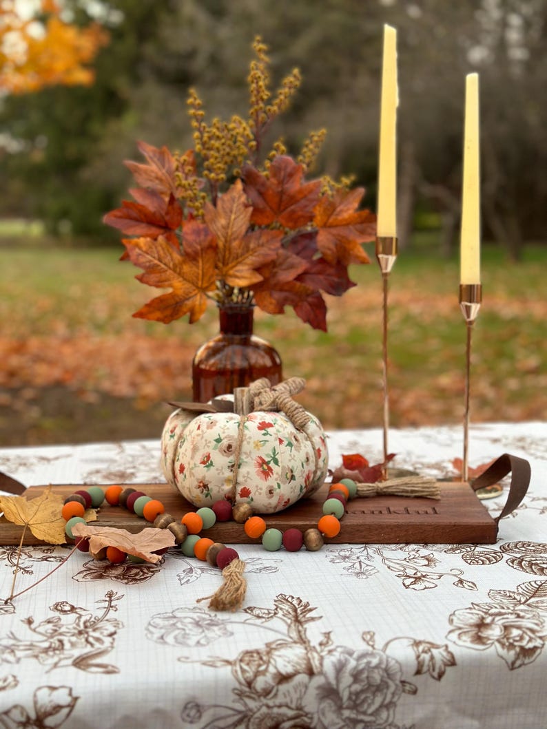 May include: A fall-themed table setting featuring a fabric pumpkin with floral patterns, a wooden tray, and a beaded garland. Two slender candles stand in copper holders, and a vase of autumn leaves adds to the seasonal decor.