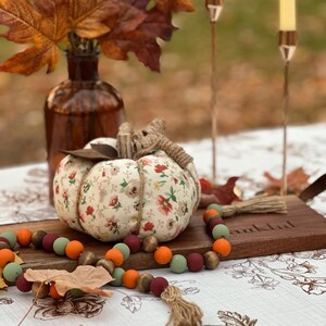 May include: A decorative fall-themed arrangement featuring a fabric pumpkin with floral patterns, a wooden bead garland, and a vase of autumn leaves. Two candle holders with lit candles are in the background. The arrangement is on a floral patterned tablecloth.