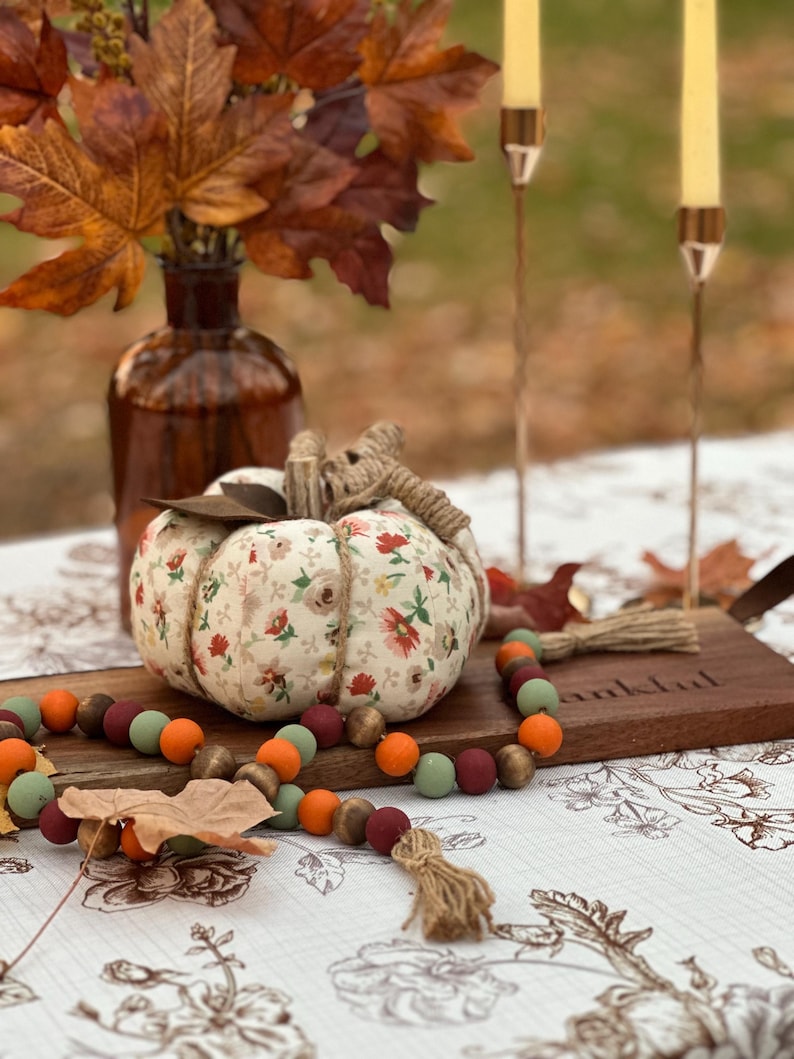 May include: A decorative fall-themed arrangement featuring a fabric pumpkin with floral patterns, a wooden bead garland, and a vase of autumn leaves. Two candle holders with lit candles are in the background. The arrangement is on a floral patterned tablecloth.