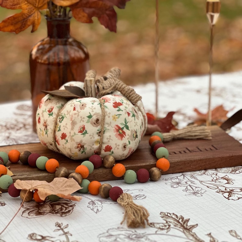 May include: A fabric pumpkin with a floral pattern, twine stem, and leaf accents sits on a wooden board. A wooden bead garland in fall colors and a brown glass bottle with fall leaves complete the autumn-themed display.
