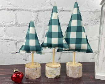 May include: Three decorative Christmas trees with teal and white plaid fabric, set on birch wood bases. Each tree has a wooden dowel trunk and a scalloped edge. A red heart-shaped ornament sits nearby.