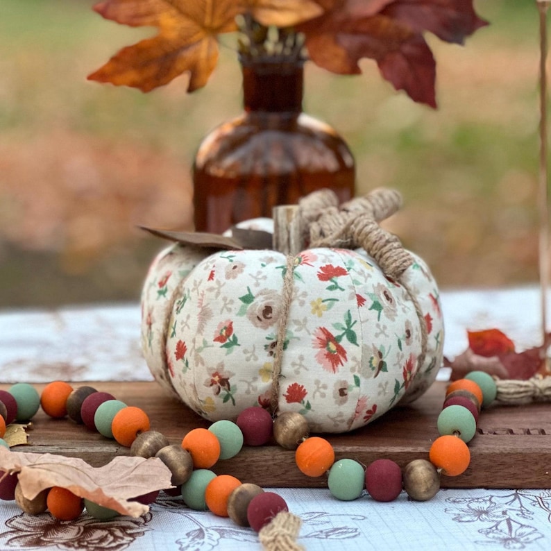 May include: A fabric pumpkin with a floral pattern, twine stem, and a wooden leaf accent. A wooden bead garland in fall colors and a brown glass vase with autumn leaves complete the fall-themed decor.