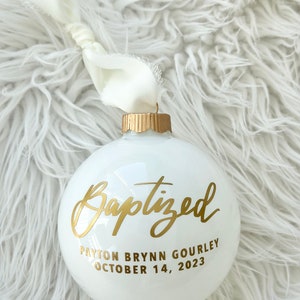 May include: A white glass Christmas ornament with gold lettering that reads "Baptized", the name "Payton Brynn Gourley", and the date "October 14, 2023". The ornament has a gold cap and a white ribbon.
