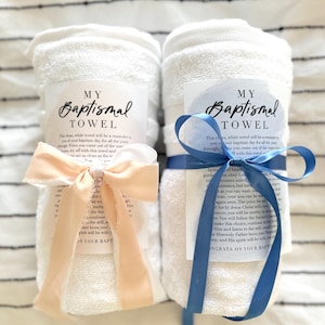 May include: Two rolled white baptismal towels, each tied with a ribbon and a tag that reads "MY BAPTISMAL TOWEL." One ribbon is peach, the other is blue. The towels are on a striped background.