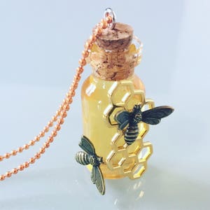 May include: A gold necklace with a small glass bottle filled with a honey-colored liquid. The bottle has a cork stopper and is decorated with a gold honeycomb and two bronze bee charms.