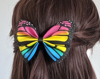 Puede incluir: Un lazo para el pelo con forma de mariposa arcoíris con una banda elástica negra. El lazo está hecho de tela y tiene una gama de colores amarillo, naranja, rojo, rosa, azul y morado.