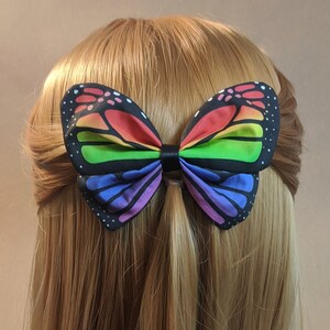 May include: A rainbow butterfly hair bow with black accents. The bow is made of fabric and has a rainbow pattern with black spots.