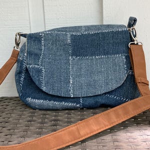 May include: A denim patchwork crossbody bag with a brown leather strap. The bag features a curved flap and is constructed from various denim squares stitched together with white thread. The adjustable strap is a light brown color.