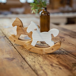 May include: Two wooden rocking horse toys. One is natural wood, the other is painted white. Both are on a wooden surface. A brown glass bottle and greenery are in the background.