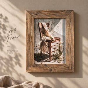 May include: A rectangular A4 picture frame made of brown reclaimed wood. The frame contains a print of a wooden chair draped with a light-coloured fabric, surrounded by small white flowers against a textured wall. The text A4 Frame Reclaimed Wood is visible.