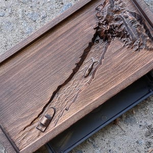May include: A rectangular, dark brown wooden tray with a carved topographical design. The tray has a raised border and a removable, dark grey base. The carved design depicts a landscape with a river and a boat.