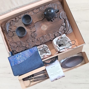 May include: A wooden tea set with a carved tray featuring lotus flowers and fish, a black teapot, two cups, and other accessories. The set includes a blue patterned pouch, small drawstring bags, and a wooden serving dish, all presented in a box.