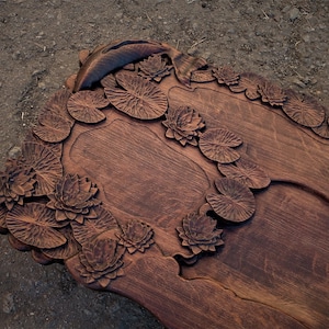 May include: A wooden serving tray carved with a detailed design of lily pads and flowers. The tray has a dark brown finish and features a small, circular, carved design with the Chinese character for "tea" in the center.