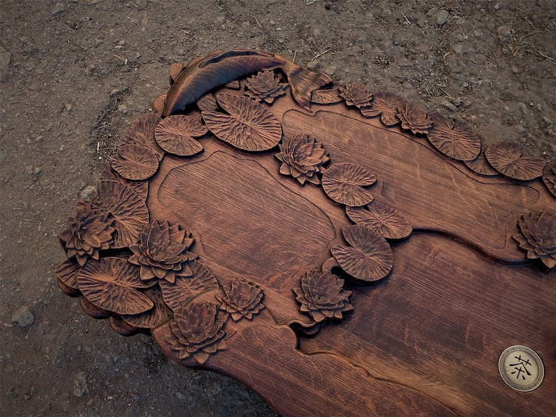 May include: A wooden serving tray carved with a detailed design of lily pads and flowers. The tray has a dark brown finish and features a small, circular, carved design with the Chinese character for "tea" in the center.