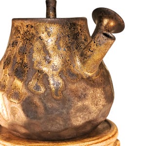 May include: A handcrafted ceramic teapot with a unique, textured surface. The teapot features a dark brown base with gold and bronze accents, a spout, and a handle. The teapot is likely used for brewing tea and is placed on a wooden base.