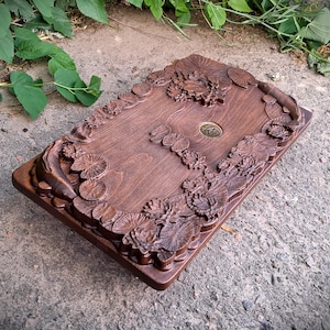 Pu&ograve; includere: Una scatola rettangolare in legno con intagli intricati di ninfee e fiori. La scatola &egrave; tinta di un colore marrone scuro e presenta un piccolo emblema metallico circolare al centro. I bordi sono anch'essi intagliati con motivi floreali.