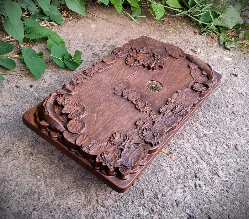 Pu&ograve; includere: Una scatola rettangolare in legno con intagli intricati di ninfee e fiori. La scatola &egrave; tinta di un colore marrone scuro e presenta un piccolo emblema metallico circolare al centro. I bordi sono anch'essi intagliati con motivi floreali.
