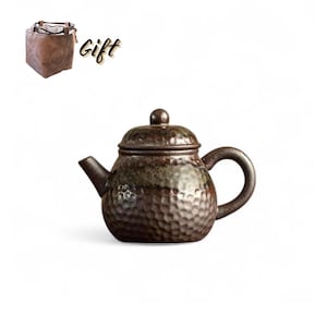 May include: A brown ceramic teapot with a textured surface and a curved handle. The teapot has a lid with a small knob and a spout. A brown leather gift bag is in the background with the word "Gift".