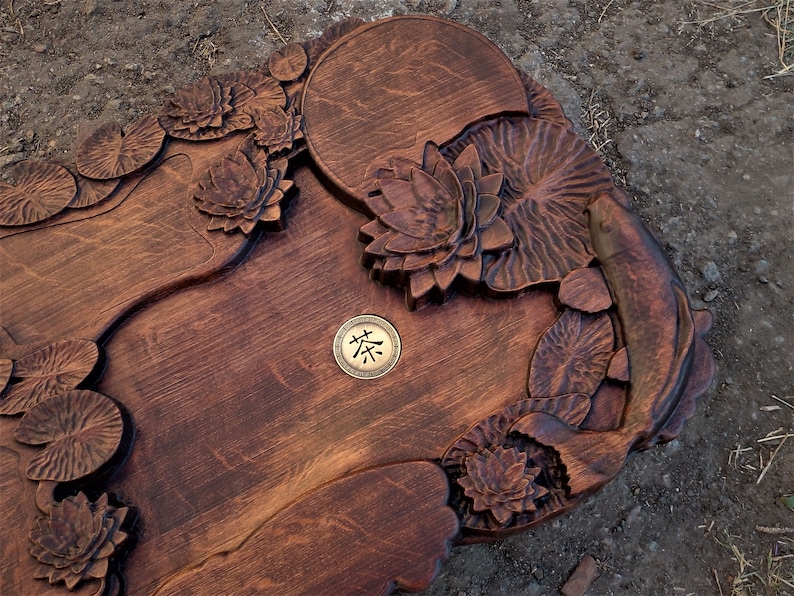 May include: A wooden tea tray with intricate carvings of lotus flowers and a fish. The tray has a round, recessed area in the center with a brass plaque that reads "茶".