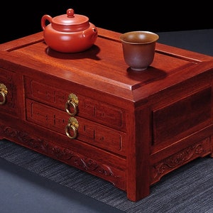 May include: A dark red wooden tea box with two drawers and intricate carvings. The box has a top surface for holding a teapot and a cup. The teapot is red and the cup is brown.