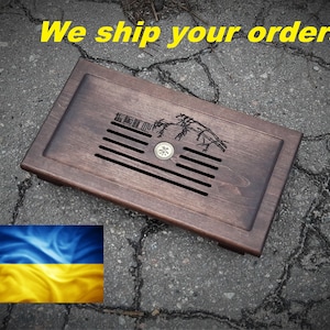 Puede incluir: Una bandeja de té de madera marrón oscuro con un diseño tallado de una rama de árbol y un pequeño desagüe de metal en el centro. La bandeja tiene un borde elevado y está sentada sobre una superficie de concreto gris. El texto "We ship your orders!" está escrito en letras amarillas sobre la bandeja.