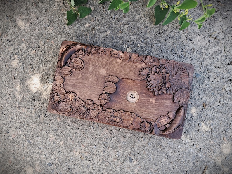 Pu&ograve; includere: Un vassoio rettangolare in legno con intagli intricati di ninfee, fiori di loto e pesci. Il vassoio &egrave; tinto di un colore marrone scuro e presenta un piccolo emblema circolare al centro. Il design &egrave; dettagliato e artistico.