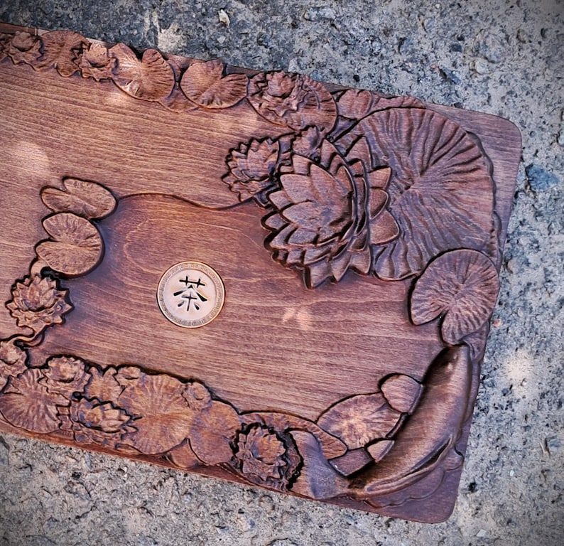 Pu&ograve; includere: Vassoio di legno marrone scuro con fiori di loto, ninfee e un pesce intagliati. Il vassoio ha un inserto circolare con caratteri asiatici. L'intaglio dettagliato crea un design naturale e artistico.