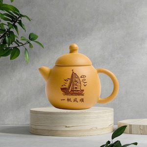 Könnte beinhalten: Eine senfgelbe Keramik-Teekanne mit Deckel und Henkel. Die Teekanne zeigt eine Segelboot-Illustration und chinesische Schriftzeichen. Sie steht auf einem Holzsockel vor grauem Hintergrund. Die Teekanne ist ein dekorativer Artikel.