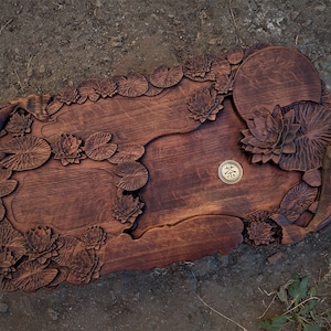 May include: A wooden serving tray carved with a lotus pond design. The tray features intricate details of lotus leaves and flowers, as well as a small round area for a teacup. The tray is stained a rich brown color.