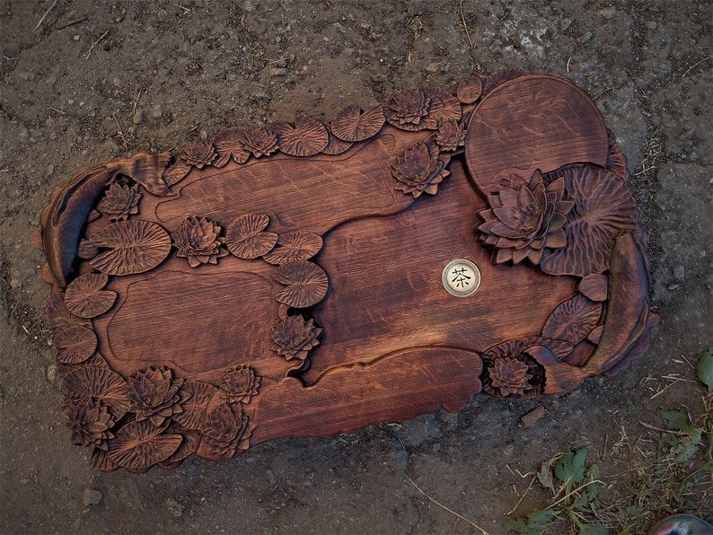 May include: A wooden serving tray carved with a lotus pond design. The tray features intricate details of lotus leaves and flowers, as well as a small round area for a teacup. The tray is stained a rich brown color.