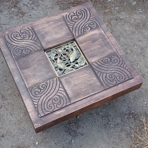 May include: A square wooden coffee table with a carved design of swirling waves. The table has a brass inlay in the centre featuring two fish and a lotus flower.