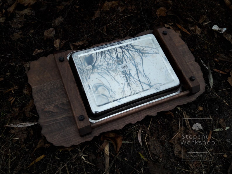 May include: A wooden serving tray with a decorative edge and a stainless steel insert. The tray is dark brown and has a natural wood grain. The insert is shiny and reflects the surrounding environment.