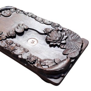Pu&ograve; includere: Vassoio da t&egrave; rettangolare in legno marrone scuro con fiori di loto intagliati, foglie di ninfea e un motivo a carpa koi. Il vassoio ha un emblema circolare centrale con caratteri asiatici. I bordi sono rialzati e la venatura del legno &egrave; visibile.