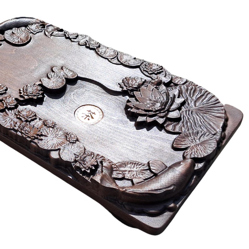 Pu&ograve; includere: Vassoio da t&egrave; rettangolare in legno marrone scuro con fiori di loto intagliati, foglie di ninfea e un motivo a carpa koi. Il vassoio ha un emblema circolare centrale con caratteri asiatici. I bordi sono rialzati e la venatura del legno &egrave; visibile.