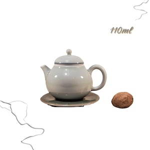 Puede incluir: Una tetera de cerámica azul claro con tapa y asa, sobre un platillo a juego. El texto "110ml" es visible. Una nuez marrón está al lado de la tetera. Capacidad de 110 ml.