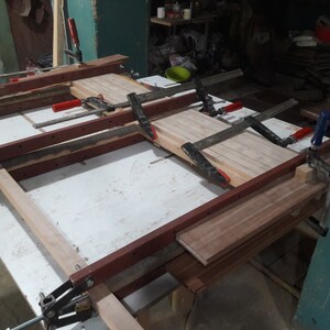 May include: A wooden table top being assembled with clamps. The table top is made of multiple pieces of wood that are being glued together. The clamps are holding the pieces of wood together while the glue dries.