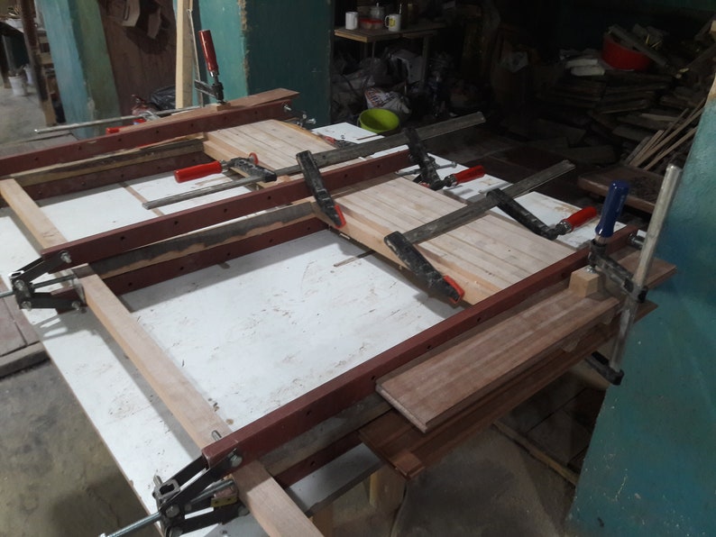 May include: A wooden table top being assembled with clamps. The table top is made of multiple pieces of wood that are being glued together. The clamps are holding the pieces of wood together while the glue dries.