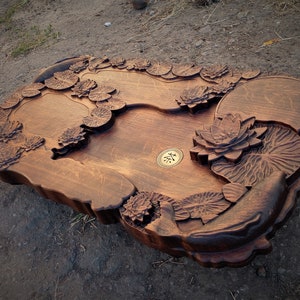 May include: A dark brown wooden tea tray with a carved lotus flower design. The tray has three separate compartments and a small brass circle in the center with the Chinese character for "tea" inscribed on it.