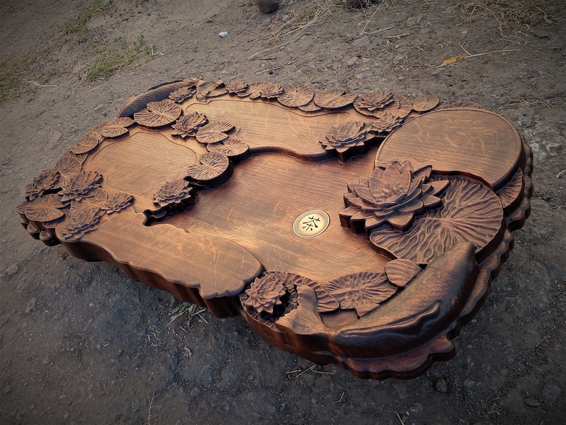May include: A dark brown wooden tea tray with a carved lotus flower design. The tray has three separate compartments and a small brass circle in the center with the Chinese character for "tea" inscribed on it.
