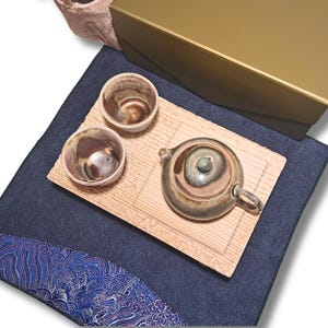 May include: A Japanese tea set featuring a silver-toned teapot and two matching cups, presented on a wooden tray. The cups and teapot have a brown and green glaze. The set is placed on a dark blue cloth with a decorative blue and white patterned edge.